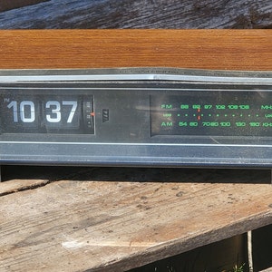 May include: A vintage wood-grain clock radio with a digital display showing the time 10:37 PM. The radio has a black face with a green digital display and a silver trim. The radio has a wood-grain top and a black base.