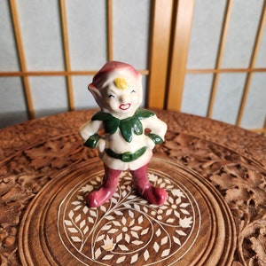 Könnte beinhalten: Eine Keramikfigur eines Elfen mit einem grünen Hut und roten Schuhen. Der Elf steht mit den Händen in den Hüften und hat einen schelmischen Gesichtsausdruck.