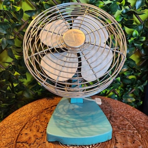 May include: A vintage teal electric fan with a chrome cage and blades. The fan has a round cage, four white blades, and a teal base. The fan is sitting on a wooden surface.