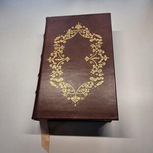 May include: A dark brown leather-bound book with a gold decorative design on the front cover. The book has a cream-coloured ribbon bookmark.