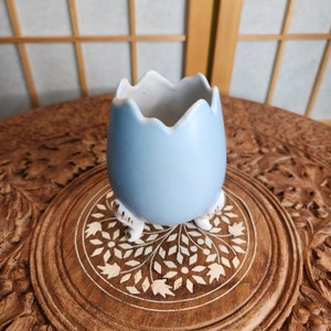 May include: A pale blue ceramic egg-shaped planter with a white interior and three small feet. The planter is on a carved wooden surface with a floral design. The egg has a jagged top edge.
