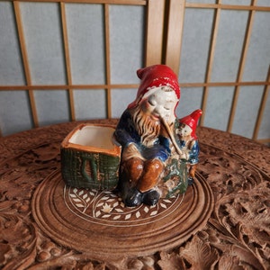 Könnte beinhalten: Ein Keramik-Pflanztopf in Form eines Gnoms mit roter Mütze, der eine Pfeife hält und mit einem kleineren Gnom sitzt. Der Topf hat ein grünes und braunes Korbmuster und ein weißes Inneres.