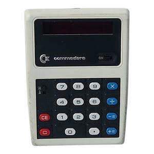 Könnte beinhalten: Ein alter Commodore-Taschenrechner in Weiß und Schwarz. Er hat eine rote Anzeige und eine Tastatur mit schwarzen und blauen Tasten. Das Commodore-Logo ist oben zu sehen.