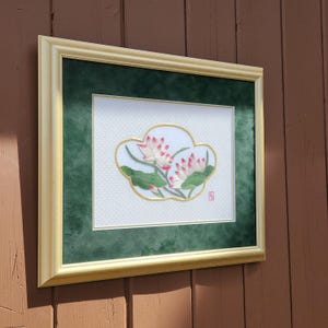 May include: Framed artwork featuring an embroidered floral design. The artwork showcases two pink and white lotus flowers with green leaves, set against a white background. The design is framed with a gold border and a green mat, encased in a light-colored wooden frame.