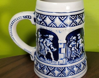 Op de afbeelding: Een witte keramische bierkan met een blauw-wit ontwerp. De kan heeft een scène met mensen in traditionele kleding.