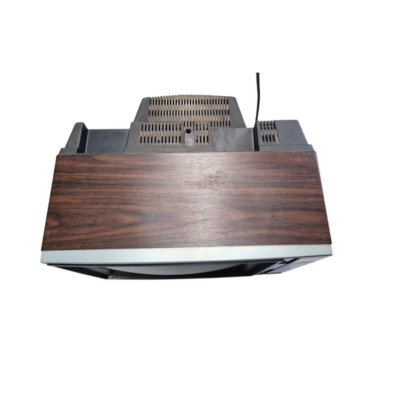 May include: A vintage television set with a dark wood grain effect on the front and top. The TV has a silver trim around the screen and a black plastic top with ventilation slots and an antenna.