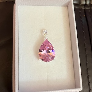 May include: A pink teardrop-shaped pendant with a silver bail, set in a white jewelry box. The pendant is faceted, catching light to create a sparkling effect. The box is open, revealing the jewelry against a white interior.