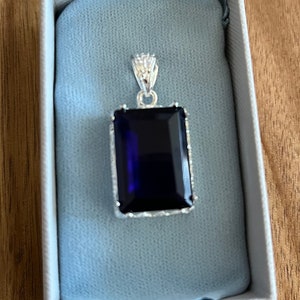 May include: A silver pendant with a dark blue rectangular gemstone. The pendant is in a white box with a blue velvet lining.