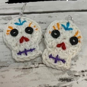 Può includere: Un paio di orecchini a forma di teschio di zucchero in crochet bianco con ricami colorati e occhi a bottone neri. Gli orecchini sono su uno sfondo bianco. Solo il modello!!