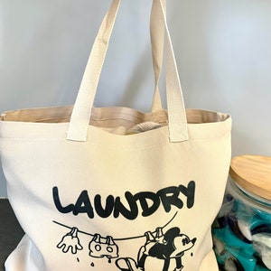 May include: A beige canvas tote bag with black text that reads "LAUNDRY". The bag features a cartoon illustration of a character hanging laundry on a clothesline. The bag has two beige handles.