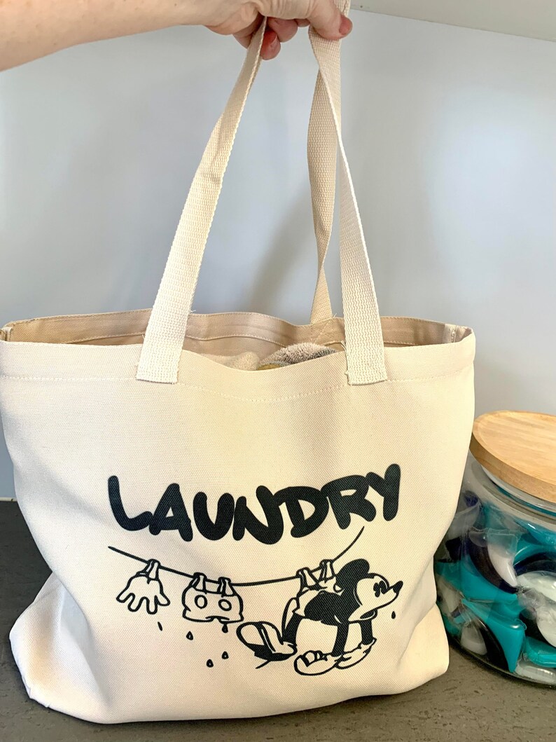 May include: A beige canvas tote bag with black text that reads "LAUNDRY". The bag features a cartoon illustration of a character hanging laundry on a clothesline. The bag has two beige handles.