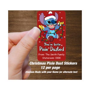 Può includere: Un adesivo rettangolare rosso con un personaggio dei cartoni animati con un cappello di Babbo Natale e luci natalizie. L'adesivo dice "You've been Pixie Dusted" e "From: The Smith Family, Stateroom: 1000". È etichettato "Christmas Pixie Dust Stickers 12 per page".