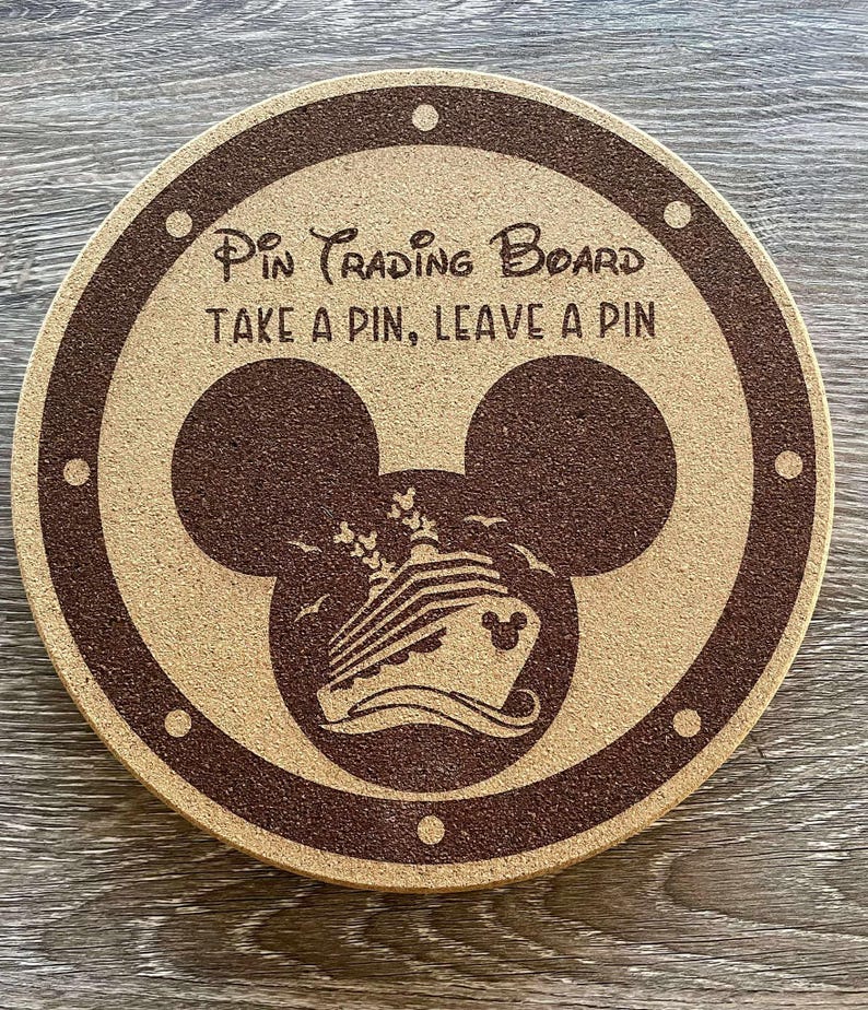 K&ouml;nnte beinhalten: Rundes Pin-Trading-Board mit braunem Rand und hellbraunem Zentrum. Das Zentrum zeigt eine Mickey-Mouse-Kopfform mit einer Kreuzfahrt-Schiff-Illustration. Der Text lautet "Pin Trading Board, Take a Pin, Leave a Pin."