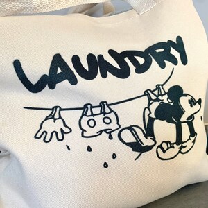 May include: Off-white canvas tote bag with a black graphic of Mickey Mouse hanging laundry on a clothesline. The word "LAUNDRY" is above the cartoon. The bag has a cream-colored handle.