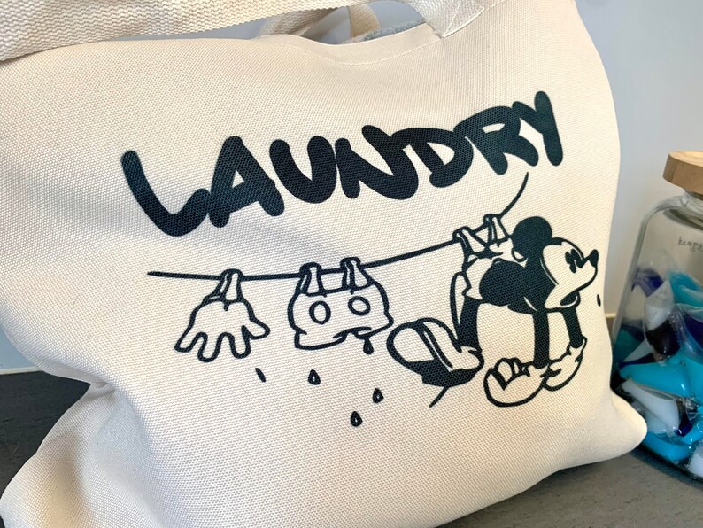 May include: Off-white canvas tote bag with a black graphic of Mickey Mouse hanging laundry on a clothesline. The word "LAUNDRY" is above the cartoon. The bag has a cream-colored handle.