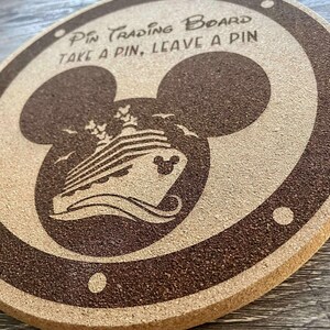 K&ouml;nnte beinhalten: Ein rundes Pin-Trading-Board aus Kork mit einem dunkelbraunen Rand und einer Mickey-Mouse-Kopf-Silhouette. Das Board zeigt ein Kreuzfahrtschiff-Design und den Text "Pin Trading Board, Take a Pin, Leave a Pin."
