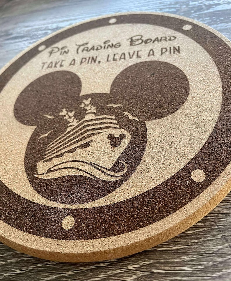 K&ouml;nnte beinhalten: Ein rundes Pin-Trading-Board aus Kork mit einem dunkelbraunen Rand und einer Mickey-Mouse-Kopf-Silhouette. Das Board zeigt ein Kreuzfahrtschiff-Design und den Text "Pin Trading Board, Take a Pin, Leave a Pin."