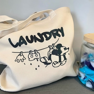 May include: A beige canvas laundry tote bag with black text that reads "LAUNDRY". The bag features a cartoon illustration of Mickey Mouse hanging laundry on a clothesline. A glass jar with a wooden lid is filled with blue and white laundry detergent pods.