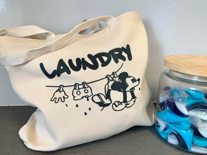May include: A beige canvas laundry tote bag with black text that reads "LAUNDRY". The bag features a cartoon illustration of Mickey Mouse hanging laundry on a clothesline. A glass jar with a wooden lid is filled with blue and white laundry detergent pods.