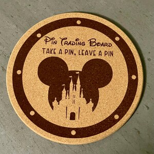 Puede incluir: Tablero redondo de corcho para intercambio de pines con borde marrón y el texto "Pin Trading Board TAKE A PIN, LEAVE A PIN". El centro presenta una silueta de orejas de Mickey Mouse y un castillo.