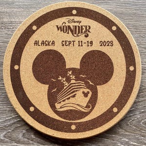 May include: Circular cork coaster with a brown border and a brown Mickey Mouse head design. The coaster features the text "Disney Wonder Alaska Sept 11-19 2023". The design includes a cruise ship and birds.