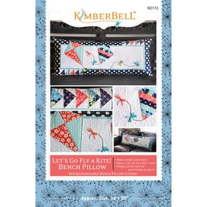 May include: A sewing pattern for a bench pillow with a kite design. The pattern includes instructions for making a pillow form and a removable cover. The cover features a colorful kite design with red, blue, and white fabric. The pattern is called "Let's Go Fly a Kite! Bench Pillow" and is by Kimberbell. The pillow is approximately 16 inches by 38 inches.