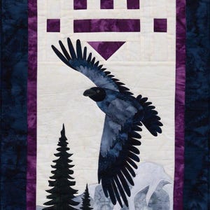 May include: A quilted wall hanging featuring a soaring raven with outstretched wings against a mountain scene. The artwork includes evergreen trees, geometric shapes, and a purple border on a cream background. The background is a dark blue fabric.