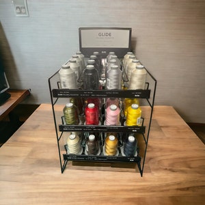 May include: A black metal display stand with two shelves holding spools of Glide Trilobal Polyester thread in various colors. The top shelf has a sign that reads "Glide Trilobal Polyester".
