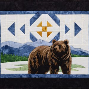 May include: A quilted wall hanging featuring a brown bear in a snowy landscape with mountains and trees. The bear is the focal point, set against a backdrop of blue and white geometric shapes and a blue border. The quilt is framed with a black border.