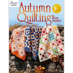 May include: The cover of "Autumn Quilting" by Annie's features three colorful quilts with fall-themed patterns. The quilts are draped over a stone wall. A bicycle with a basket and a wooden barrel with apples are in the foreground.