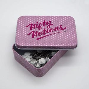 May include: A rectangular, open, lavender tin with a polka dot pattern and the words "Nifty Notions" in pink script. The tin contains pins with white flower-shaped heads.