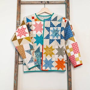 May include: A quilted jacket with a patchwork design featuring star patterns in various colors, including pink, blue, yellow, and orange. The jacket has a teal trim and is hanging on a wooden hanger.