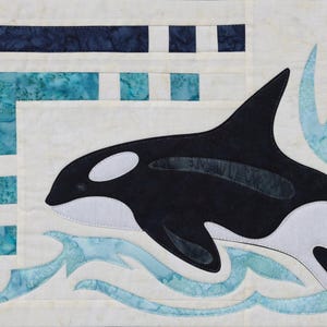 May include: A fabric art piece featuring a black and white orca whale leaping through light blue waves. The background is a cream color with a geometric design in shades of blue. The orca has white patches and a tall dorsal fin.