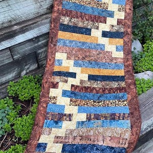 May include: A long, rectangular table runner with a geometric pattern of brown, blue, and yellow stripes. The runner is made of fabric and has a quilted texture.