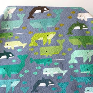 May include: A quilted fabric featuring a repeating pattern of various whales and small fish in shades of blue, green, and white on a gray background. The textile has a continuous wavy stitched texture throughout the entire surface area.