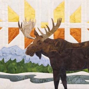 May include: A fabric art piece featuring a moose in a winter landscape. The moose is brown with large antlers, set against a backdrop of mountains, a river, and a stylized sunset in shades of yellow and orange.