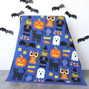 May include: A blue Halloween-themed quilt featuring repeating patterns of pumpkins, black cats, owls, ghosts, witches, spiders, bats, candy corn, and stars. The quilt is displayed on a metal rack with paper bats in the background.