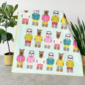 May include: A light blue quilt featuring a repeating pattern of cartoon sloths and bears wearing colorful shirts and shorts. The quilt has a yellow border and is displayed on a light gray surface.