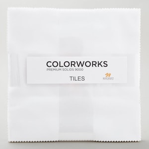 May include: A stack of white fabric squares with the text "COLORWORKS PREMIUM SOLIDS 9000 TILES" on a white label. The fabric has a subtle texture and a scalloped edge. The Northcott logo is also visible.