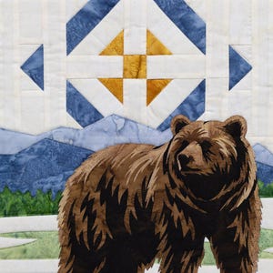 May include: A quilted textile featuring a brown bear in a mountain landscape. The bear is the focal point, set against a backdrop of blue mountains, green trees, and geometric shapes in blue and gold. The quilt has a white background.
