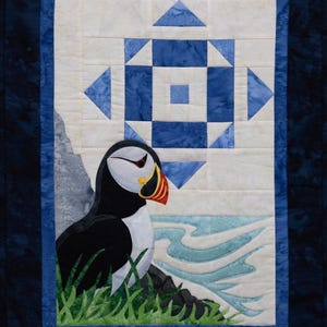 May include: A quilted wall hanging featuring a puffin bird. The bird is black, white, and orange, perched on a rock with green grass. The background has a blue and white geometric design and a blue border.