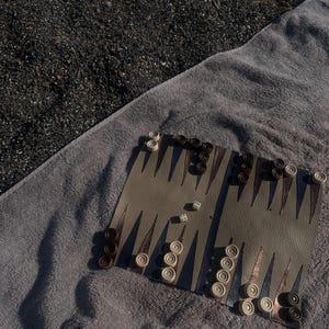 May include: A backgammon board with white and brown checkers and two dice on a gray towel. The board is tan with brown and white triangular points. The checkers are arranged on the board, ready for play. The towel is on a dark, textured surface.