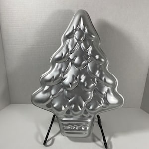 Puede incluir: Un molde para pastel plateado con forma de árbol de Navidad. El molde tiene un diseño detallado de un abeto con una base en forma de cesta. El molde es de metal y se exhibe sobre un soporte de metal negro. Ideal para hornear en las fiestas.