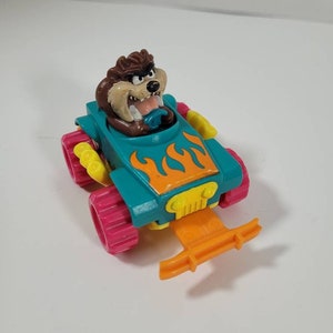 May include: A teal and orange toy car with flames on the side. The car has pink wheels and a yellow steering wheel. A brown cartoon Tasmanian Devil is driving the car.