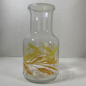 May include: A clear glass jug with a wide base and a narrow neck. The jug is decorated with a gold wheat pattern. The jug is empty and sits on a white surface.