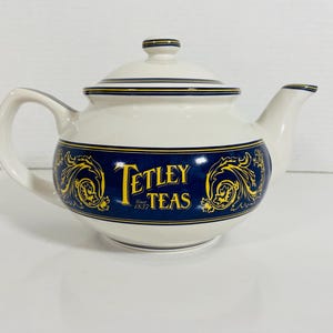 May include: Cream-coloured ceramic teapot with a lid, handle, and spout. It has a navy blue band with gold floral patterns and the text "Tetley Teas Since 1837". A traditional tea pot.
