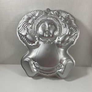May include: A silver metal cake pan shaped like a Cabbage Patch Kid doll. The doll is wearing a dress and has pigtails with bows.