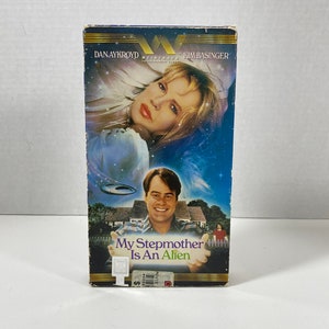 May include: VHS video cassette cover featuring Dan Aykroyd and Kim Basinger in the film "My Stepmother Is An Alien". The cover has a blue and black background with stars and a flying saucer.