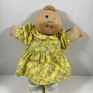May include: A Cabbage Patch Kid doll wearing a yellow dress with a floral pattern. The doll has blonde hair, green eyes, and a rosy complexion.