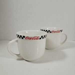 May include: Two white ceramic mugs with black and white chequered bands around the top. Both mugs have the Coca-Cola logo in red lettering.